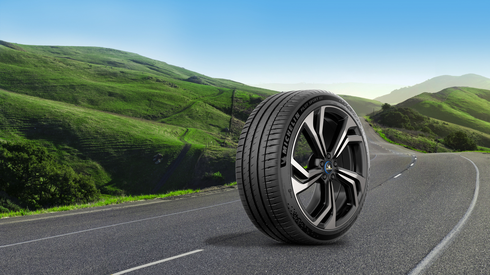 Immagine banner Michelin ruota completa di pneumatici MICHELIN Pilot Sport EV su sfondo strada montana con prati verdi